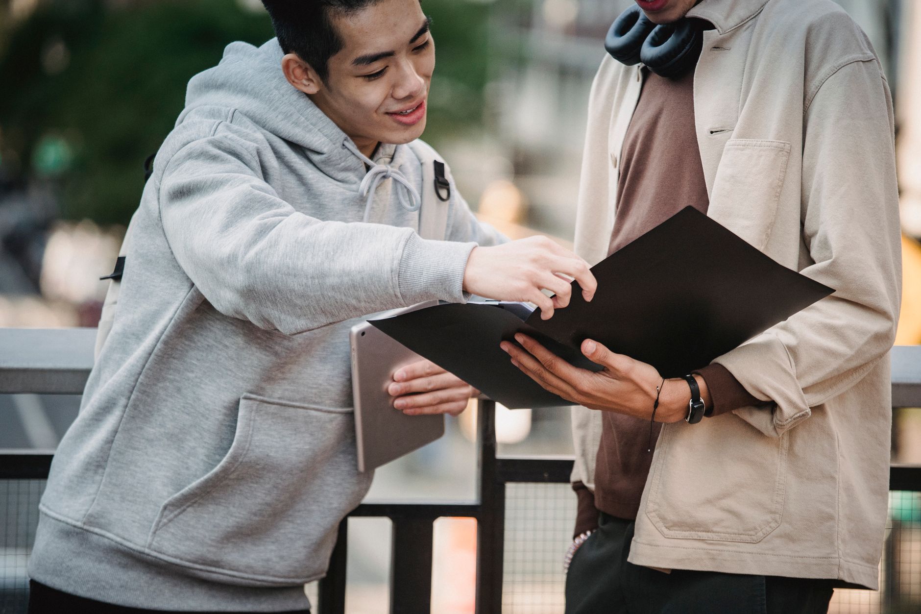 Two people outdoors looking at a black folder, one pointing at it while holding a laptop under his arm.