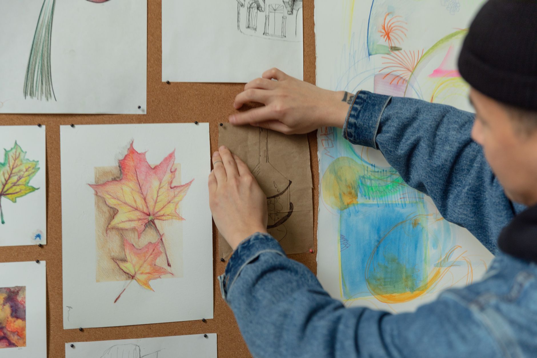 A person in a black beanie and denim jacket pinning a brown paper drawing to a corkboard covered with colorful artwork.